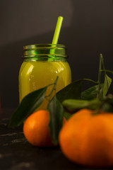 Freshly squeezed orange juice in a mason jar and fresh oranges fruit with green wet leaves on a black shabby background.
