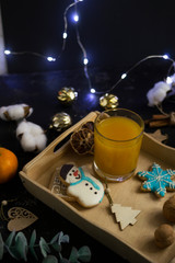Christmas cookies in the shape of a house, a snowman and stars lie in the gap with fresh orange juice in a glass of farm-style drink. Around nuts, cinnamon, decor. Black background.
