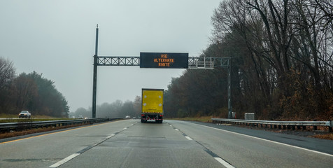 Electric warning sign over yellow box truck on highway advising drivers to use an alternate route...