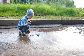 A three-year-old white boy in a blue hat, jeans and sandals plays on a cool summer day with an outdated remote control car.