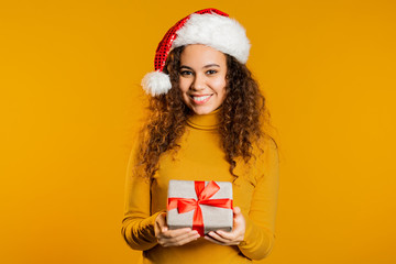 Young pretty african woman smiling and holding gift box on yellow studio background. Girl with curly hairstyle in sweater. Christmas mood.