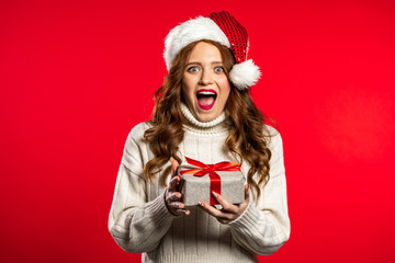 Surprised pretty woman smiling and holding gift box on red studio background. Girl with beautiful hairstyle in white sweater and Santa hat. Christmas mood.