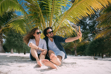 A pair of lovers take a selfie under a palm tree on a tropical beach