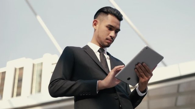 Attractive asian businessman in a black suit using his tablet digital browsing financial, scrolling social media while standing outside on the street near a big office building urban.