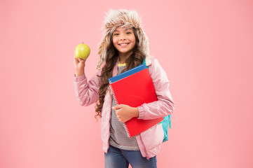 tastes great. schoolgirl with books after lesson. school break. small happy girl earflap hat eat apple. winter holiday and vacation. teen kid pink background. pupil daily life. Modern education