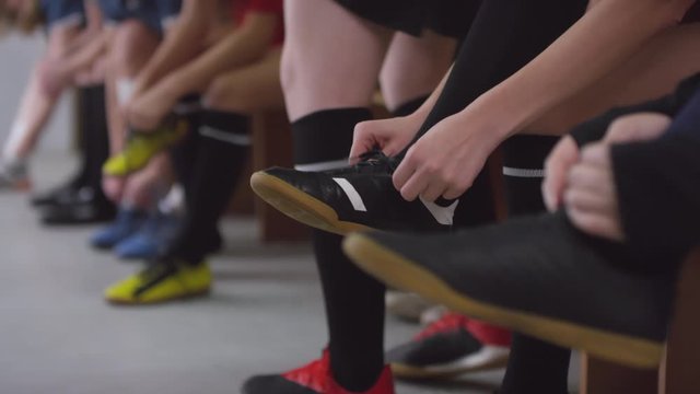 Close up selective focus shot of legs of female soccer athletes sitting on bench and tying shoelaces while preparing for training in locker room