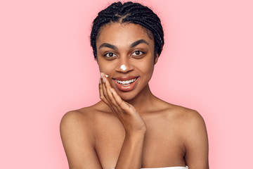 Beauty Concept. Young african woman isolated on pink applying cream on face smiling surprised