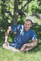 Naklejka na ściany i meble Man in blue t-shirt sitting cross-legged on grass and smiling in the garden on blurred greenery background on a sunny summer day. Vertical orientation. Toned