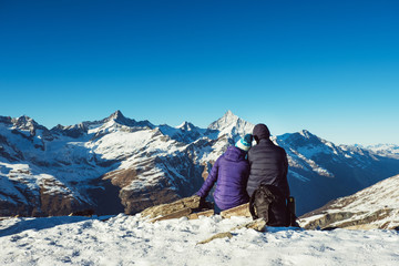 Happy travelers couple man and woman trekking in mountains, enjoy beautiful view. Explorers hiking on snowy hills, travel in Swiss Alps, Switzerland. Romantic hikers sitting outdoors on nature.