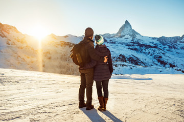 Happy travelers couple man and woman trekking in Swiss Alps, enjoy beautiful sunset view of Matterhorn mountain peak. Explorers hiking, travel in Switzerland. Romantic holidays outdoors on nature.