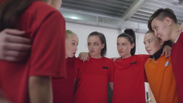 Team of young female soccer athletes in sports uniform standing in circle, embracing and listening to leader while getting ready for match in locker room