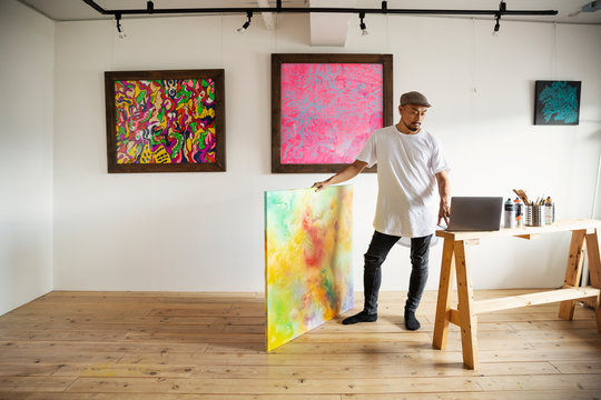 Japanese man standing in art gallery, holding abstract artwork, looking at laptop computer.