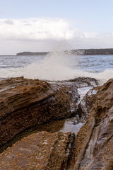 Look out. Beach with the wave of the sea hitting the rocks and splashing water everywhere. Rock Formation. Transparent blue water and shining sun.