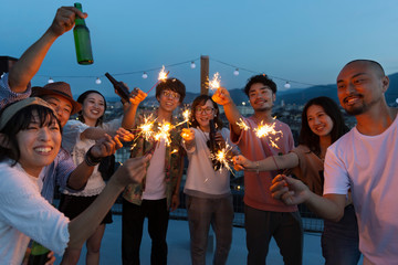 Group of young Japanese men and women with sparklers on a rooftop in an urban setting.