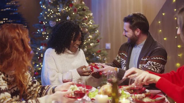 Nice-looking carefree young people celebrating Christmas Eve in company. Multi-ethnic romantic couple exchanging gifts at table having fun celebration reunion. Merry Christmas.
