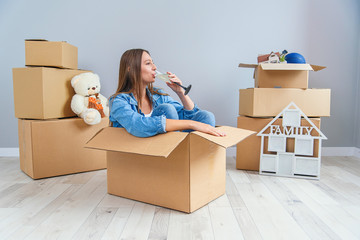 Happy woman drinks champagne from a glass while sitting inside a cardboard box in new apartment.