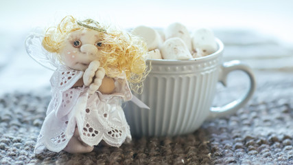 angel in white lace dress with hands folded in prayer. homemade sock toy with fluffy red hair in background of window. cup of marshmallows on knitted napkin.