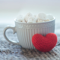 Fototapeta na wymiar A big crochet red heart next to a cup of cocoa. Cappuccino in a gray cup against a white background.
