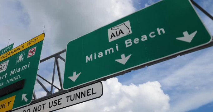A view driving under a Miami road sign on a sunny day. Miami Beach A1A North and Port Miami via Tunnel and Route 887 are shown.  	