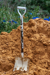 Newcastle, Australia - December 10, 2009: Closeup of white and rusty shovel standing straight in heap of orange-brown sand. Green foliage and blue flowers in back.