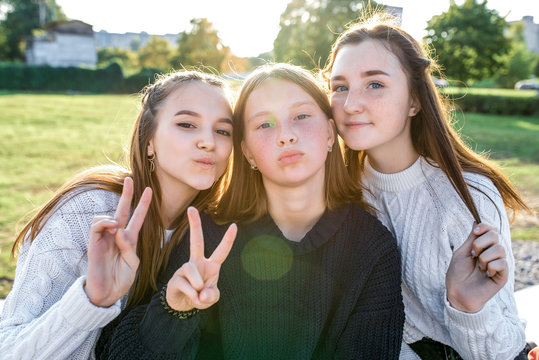 Girls girlfriends teens three teenagers, are photographed on camera, fingers hand gesture victory everyone hello, autumn outdoors in the park. Casual warm sweater clothes. Portraits close-up.