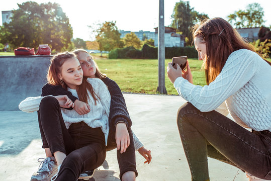 Girls girlfriends three teenagers, take pictures on phone, sit on skateboard ride, autumn is outdoors in park. Casual warm sweater clothes. Rest after lessons at school and college.