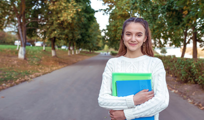 Happy teenager girl smiling cheerful joyful, stands summer park autumn trees background, free space for copy text. In hands notebook textbooks notes, preparation school college. Warm clothes sweater.
