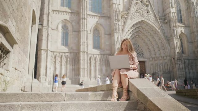 Laughing woman working with laptop near cathedral. Smiling girl getting message