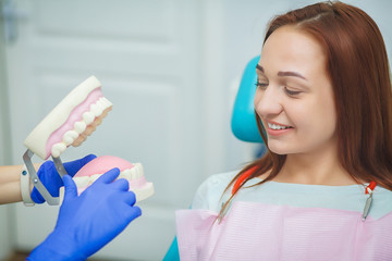 Young dentist holding a plastic model of teeth and explaining the procedure of new dental implants to her patient sitting in dental chair at stomatology