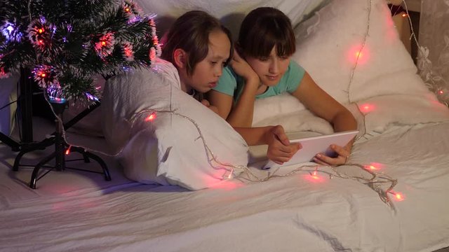 children on Christmas evening play on tablet, in children's room on couch garlands. sisters with tablet play in a room decorated with colorful Christmas tree. concept of a family Christmas.