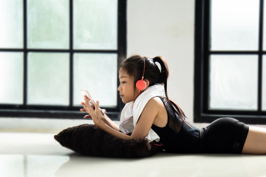 Asian little adorable schoolgirl or happy cute girl in dancing suit lie down on the floor, relaxation with colorful headphone and smartphone after practicing or training jazz dance in studio classroom
