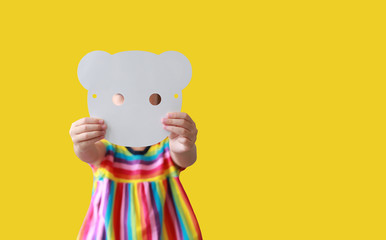 Cute little child girl showing blank white animal paper mask fronting her face isolated on yellow background with copy space. Idea and concept for kid dressed up playing animal face.