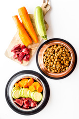 Pet feed ingredients. Raw meat and fresh vegetables near bowl with dry feed on white background top-down