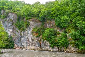 A steep rock cliff with a green forest over a mountain river on a cloudy day.