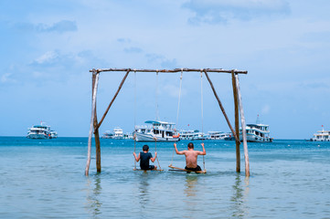 Family, father and son are spending their holidays, swinging on the swing in the sea next to the Bai Sao beach on Phu Quoc island, Vietnam. Sunny day, boats on the background.
