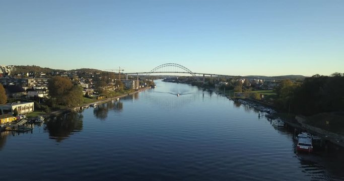 Aerial Drone following river through Fredrikstad Norway with boat approaching and large bridge in background during sunrise