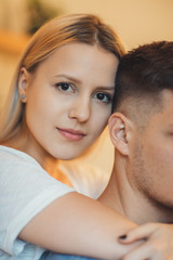 Close up portrait of beautiful young blonde woman looking at camera smiling while embracing her boyfriend's neck from back.