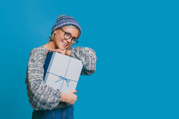 Lovely young blonde woman dressed in blue clothes looking at camera laughing while holding a blue gift box against blue background.