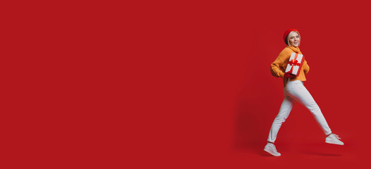 Full-length portrait of a charming young blonde woman walking while holding a red gift box looking at camera isolated on red background.