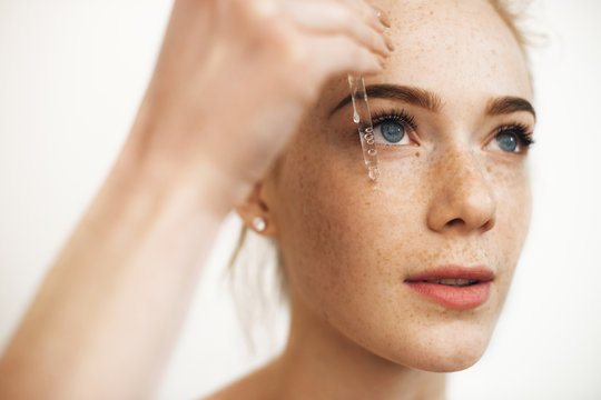 Close up portrait of a charming red haired woman with freckles applying hyaluronic serum on her face isolated on white.