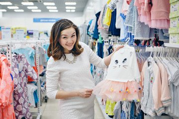 a pregnant woman in a light dress and stockings considers children's clothes in the shop. Healthy lifestyle concept, IVF fashion for pregnant women, children