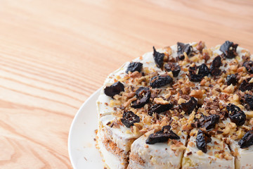 Vanilla sponge cake biscuit with prune, walnuts and frosting served on a white plate, on light wooden background table