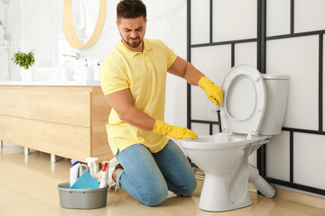 Young man feeling disgust while cleaning toilet bowl in bathroom