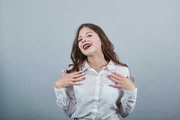 Cheerful attractive caucasian woman over isolated gray background holding hands on chest, smiling, looking aside, wearing casual white shirt