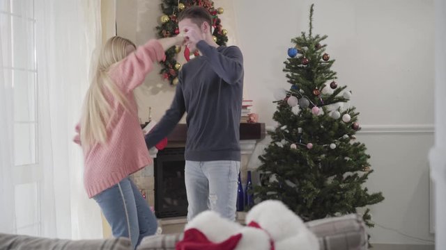 Young Caucasian man and woman dancing in front of Christmas tree at home. Positive couple spending New Year's eve together. Holidays, celebration, happiness.