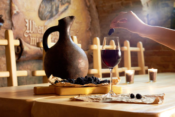 Woman hand with mulberry, glass of red wine, clay jug on wooden table in a wine cellar restaurant