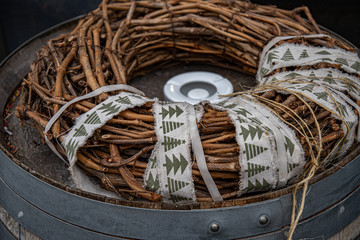 Rustic Christmas wreath made from woven grapevine branches and wrapped by old torn white ribbon with primitive pattern of Christmas tree silhouettes. Top of wooden barrel with riveted metal hoops