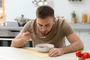Young man eating tasty vegetable soup at table in kitchen