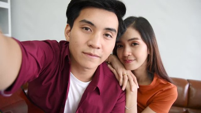 Young Asian couple lover sitting on couch and take a selfie together in modern living room background