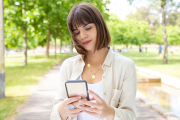 Content young woman using smartphone outdoors. Beautiful cheerful young woman standing and using mobile phone in park. Technology concept